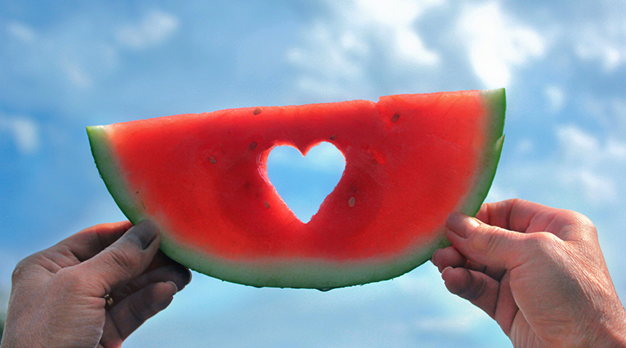 Hands holding a slice of watermelon with a heart cut out of it.