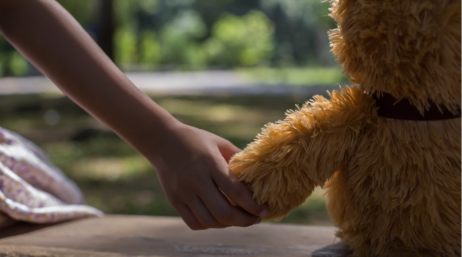 A child sits on a bench, holding her teddy bear's hand