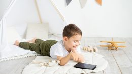 A child lies on the floor under a canvas tent in his bedroom, with a tablet in front of him