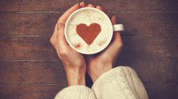 A woman holding a cup of coffee with milk foam on top and a heart made of cocoa