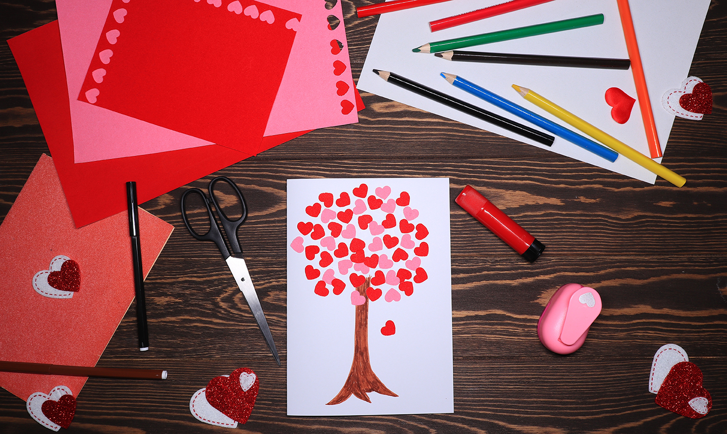 A piece of paper with a tree drawn on it and paper hearts for the leaves. In the background are craft supplies such as coloured paper, pencil crayons, scissors, and a heart punch. 
